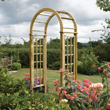 Drewniana pergola ogrodowa z łukowatym zwieńczeniem, otoczona kwitnącymi kwiatami i bujną zielenią, na tle pochmurnego nieba.